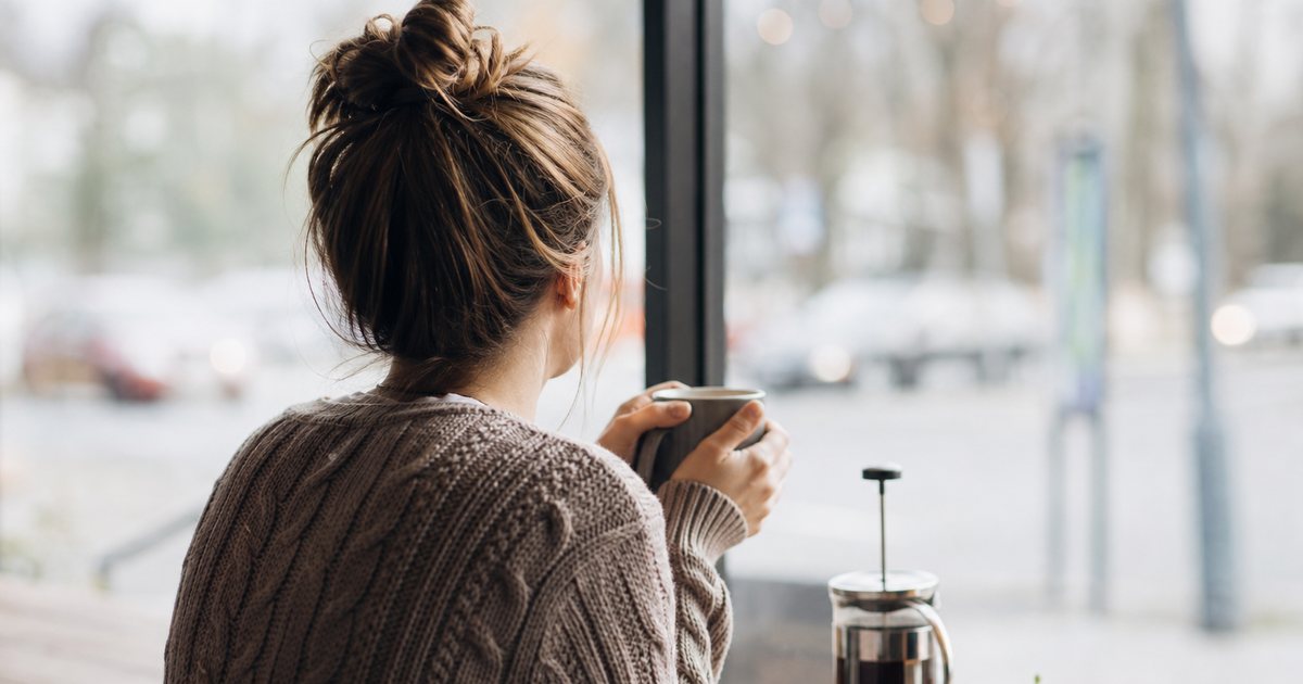 A person photographed from behind at a cafe window table, hands wrapped around a mug, blurred street outside, soft morning light, face not visible
