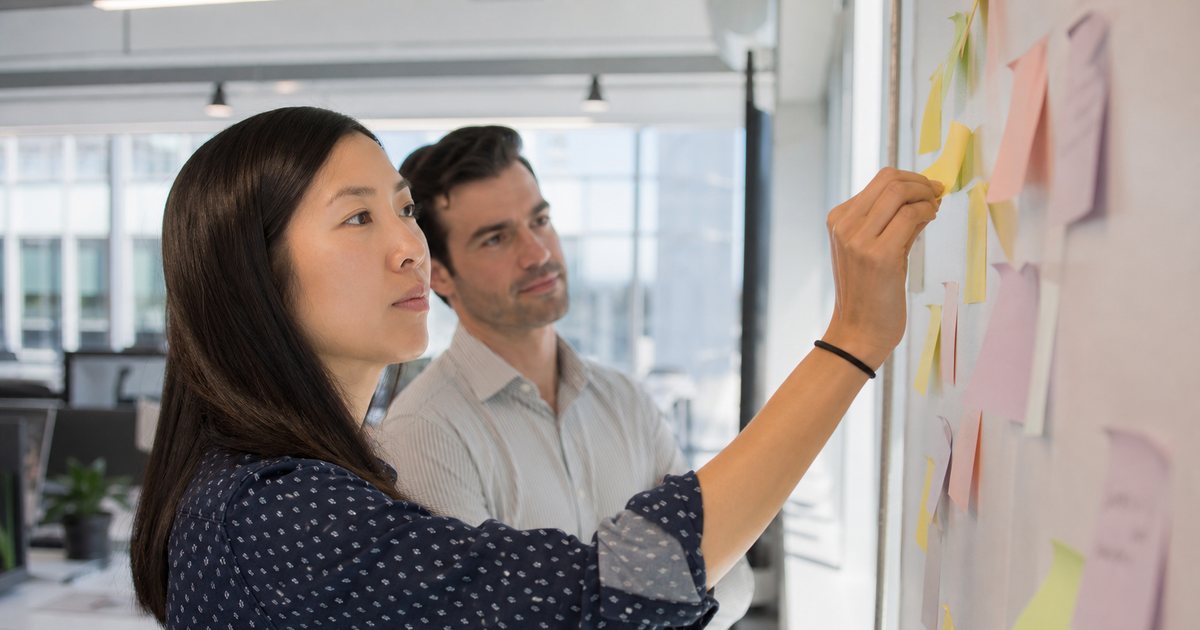 Two colleagues in a bright open plan office standing at a wall covered with blank pastel sticky notes, one reaching up to peel a faded note away, tall windows and soft morning daylight