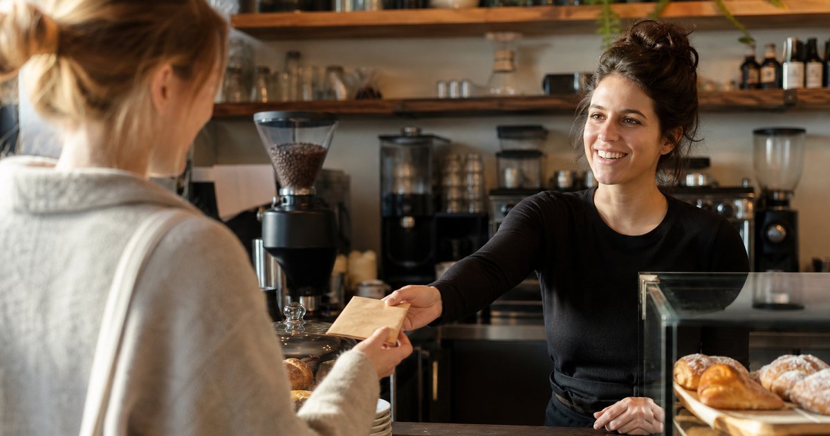 A barista at a coffee shop counter handing a small blank paper envelope to a smiling customer, pastries under glass, warm afternoon light