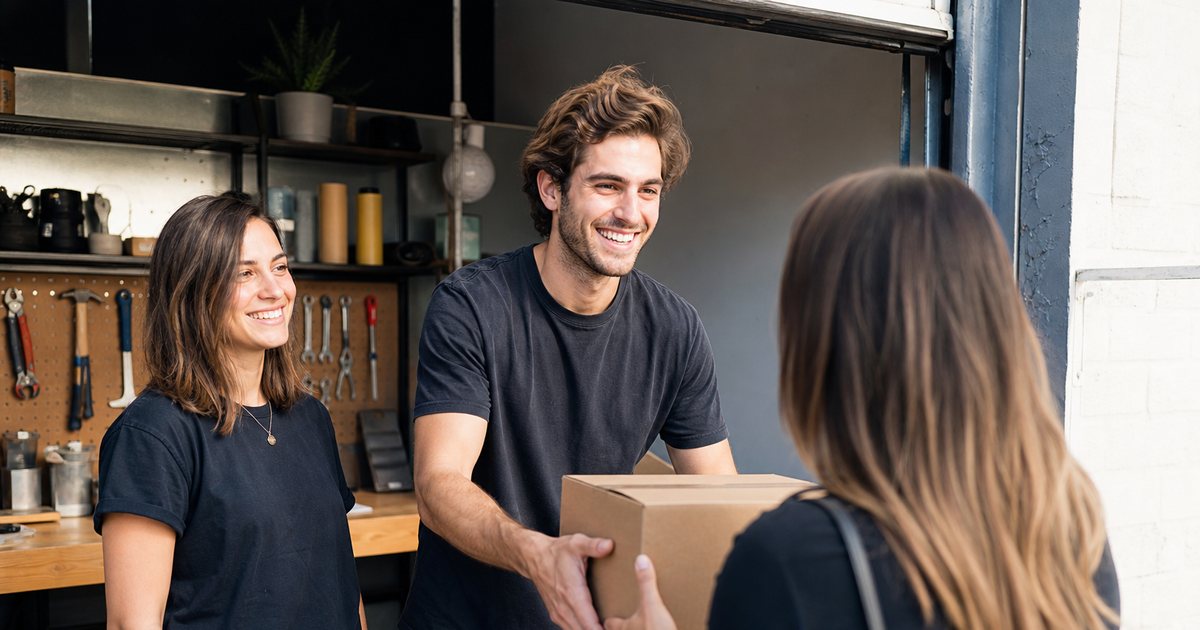 Two founders in a small garage workshop handing a freshly packed cardboard box to their first customer at the open door, sunlight, tools on a pegboard behind them